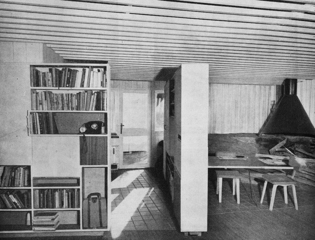 Looking into the kitchen with a fixed storage unit on the left. The central storage unit is hinged at the rear column with a fixed castor at the nearest end, enabling the wall to be moved left or right to maximise spaces as and when needed. The dining table is hinged at the left end and folds up into the storage wall, enabling the fireplace area to be more fully used after dinner. The curtains pull to the left revealing a stone wall behind and the large glazed wall to the right and north which lets the sun in. The glass runs right down into the soil without any sill, enhancing the continuity of indoor space with the outdoors. The original metal hood has yet to receive its render coating as in the other photo.